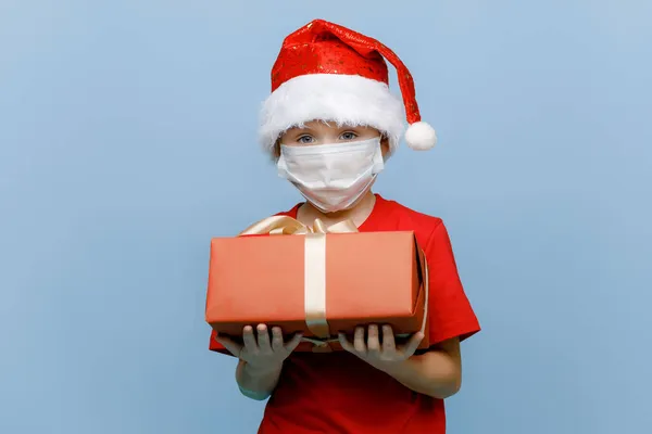 Tıbbi maske takan, Noel şapkası takan, hediye kutusunu tutan ve mavi arka planda izole edilmiş kameraya bakan mutlu küçük bir çocuğun portresi. Boşluğu kopyala