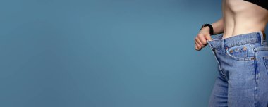 30 'larında, geniş kot pantolon ve beyaz gömlek giyen çekici, narin genç bir kadın başarılı bir kilo kaybı gösteriyor. Diyet ve kilo kaybı konsepti..