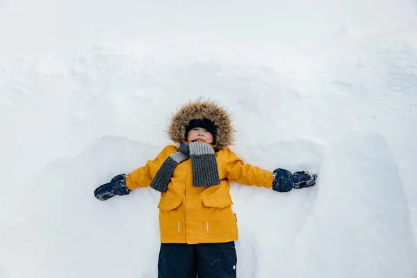 Menino bonito em um casaco laranja com um capuz de pele está se divertindo deitado na neve. Atividades ao ar livre de inverno para crianças. Vista superior — Fotografia de Stock