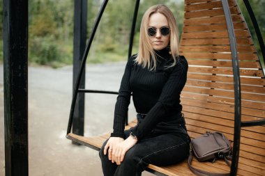 Havalı genç sarışın dışarıda sallanan yuvarlak bir sandalyede oturuyor. Model bir sandalyeye oturur ve rahatlar, kameraya bakar.