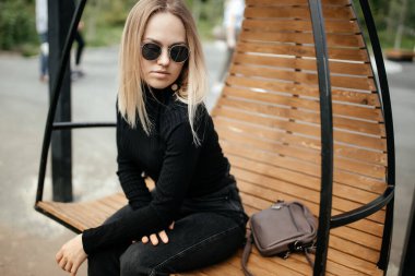 Havalı genç sarışın dışarıda sallanan yuvarlak bir sandalyede oturuyor. Model bir sandalyeye oturur ve rahatlar, kameraya bakar.