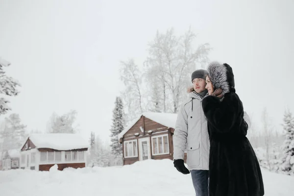 Hermosa pareja enamorada, sonriendo y mirándose, una pequeña casa de madera y nieve en el fondo — Foto de Stock