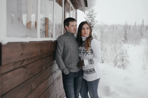Hermosa pareja enamorada abrazándose, pequeña casa de madera y nieve en el fondo — Foto de Stock