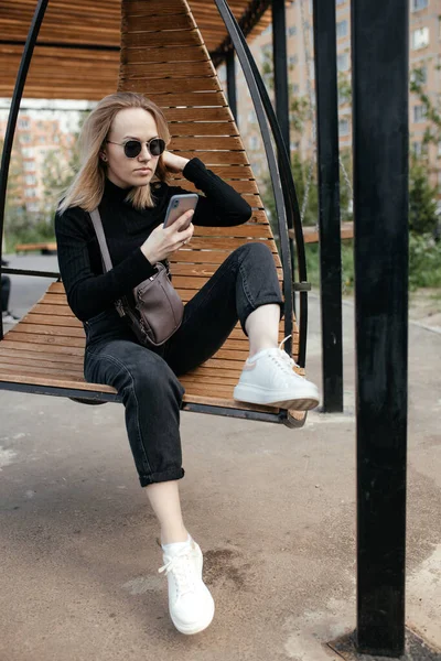 Belle femme de 30 ans avec des lunettes envoie des SMS et prend des selfies sur une caméra smartphone dans un parc, à l'extérieur. Jolie blonde souriante se repose sur un banc et utilise un téléphone portable. — Photo