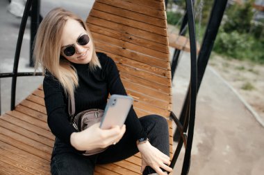 Yakın plan 30 yaşında gözlüklü güzel bir kadın parktaki bir akıllı telefon kamerasında mesaj atıyor ve selfie çekiyor. Güzel gülümseyen sarışın bankta dinleniyor ve cep telefonu kullanıyor.. 