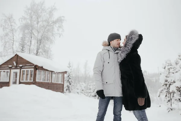 Hermosa pareja enamorada, sonriendo y mirándose, una pequeña casa de madera y nieve en el fondo — Foto de Stock