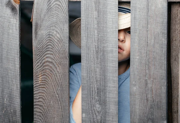 Five- to six-year-old kid in a straw hat peeps through a wooden village fence. Copy space — Stock Photo, Image