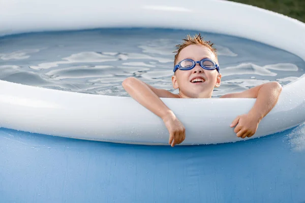 Un guapo niño de cinco años con botas de baño y cabello desaliñado se baña en una piscina inflable azul. Funny Kid juega en la piscina para niños y mira a la cámara — Foto de Stock