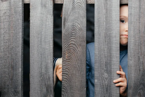 Happy smiling white boy looks out of the crack of a wooden fence. Childish curiosity. Espionage. Rural life. Child development, curiosity. The boy peered through the crack. — Stock Photo, Image
