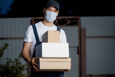 Tıbbi maskeli kurye kılığındaki erkek kurye açık havada elinde bir paket tutuyor. Kurye kavramı paketini her an teslim edebilir.. 