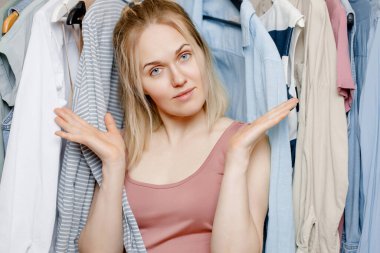 Image of a charming young Caucasian woman shrugging her shoulders, standing among fashionable clothes and feeling embarrassed because she doesn't know what to wear to a party. 