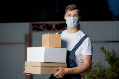 Üniformalı ve maskeli kurye elinde bir yığın kutu tutuyor. Kurye teslimat servisi açık havada paketleri elinde tutuyor.. 