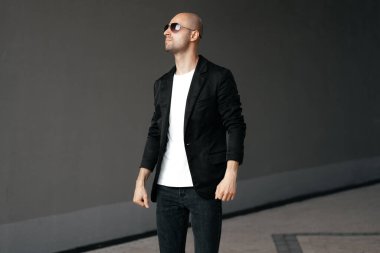  Gözlüklü genç kel bir adam ve açık havada şık kıyafetler. Gri bir arka planda yarım bir dönüşle yan yana duruyor.