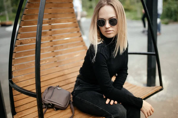 Genç, güzel, beyaz bir kız bir park bankında oturuyor. Hoş bir kadın yaşam tarzı, yumuşak odaklı bir portre. Düşünceli, sakin yüzlü, şık bir kız.