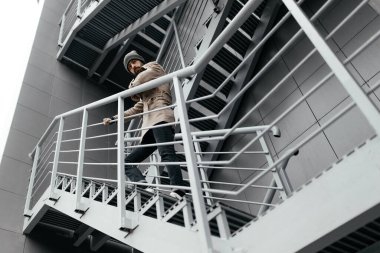 Paltolu, kot pantolonlu ve şapkalı şık genç sakallı hippi şehirdeki gri metal bir merdivende kameraya bakar. Çekici, modern adam. Sokak tarzı. sonbahar.