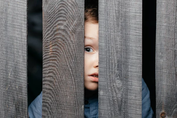 Aranyos kis fehér fiú fél szemmel átlát egy repedésen egy rusztikus fa kerítésen. — Stock Fotó