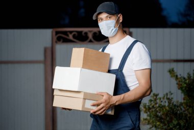 Maskeli ve üniformalı yakışıklı teslimatçı kapıda bir yığın kutu tutuyor. Elinde piyonları olan bir erkek kurye kameraya bakar.. 