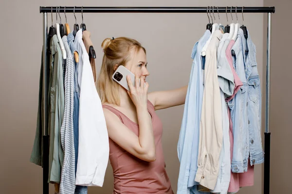Attraktiv kvinna pratar på sin smartphone, står mellan hängare med kläder. Kvinna pratar i telefon med en vän och tänker vad man ska ha på sig på ett möte med vänner. Stockbild