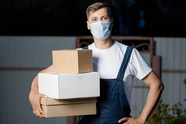 Courier homem de uniforme e máscara mantém uma pilha de caixas em suas mãos. Serviço de entrega de correio com parcelas na mão ao ar livre Imagem De Stock