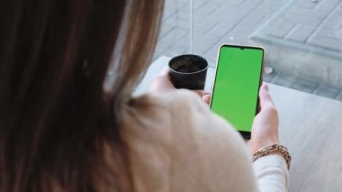 Kafede elinde yeşil ekranlı akıllı telefon tutan bir kadın var.