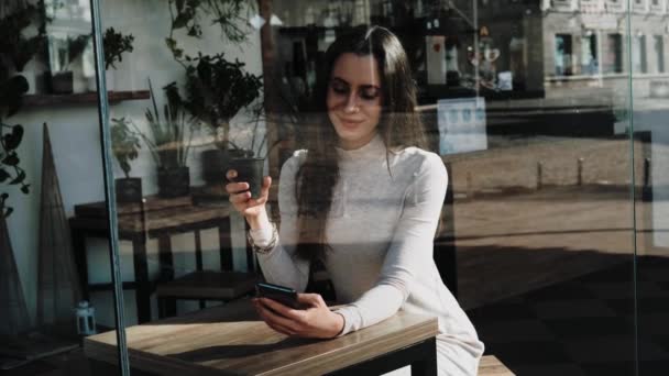 Mujer joven y bonita con un teléfono inteligente en la cafetería con una taza de café sonriendo — Vídeo de stock