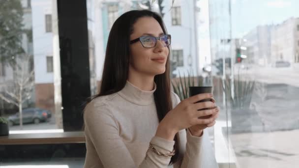 Молодая женщина в очках пьет кофе и смотрит городскую жизнь — стоковое видео