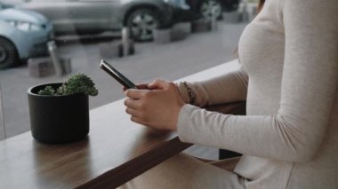 Akıllı telefon kullanan ve büyük pencerenin yanındaki kafede oturan siyah bardaktan kahve içen genç bir kadın şehir hayatını izliyor. El bilgisayarı ağır çekim orta çekim.