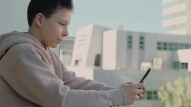 Kafkasyalı kapüşonlu genç balkonda akıllı telefon kullanarak sohbet ediyor. Şehir manzarası.