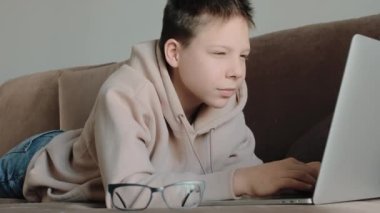 Kapüşonlu, gözlüklü, kanepede dizüstü bilgisayar kullanan beyaz bir çocuk.