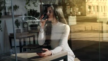 Akıllı telefonlu güzel bir kadın internette geziniyor ve bir fincan kahveyle kafeye bakıyor. Gündüz vakti orta atış. Pencereden bak. El bilgisayarı ağır çekim.