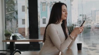 Akıllı telefon kullanan ve büyük pencerenin yanındaki kafede oturan siyah bardaktan kahve içen genç bir kadın şehir hayatını izliyor. El bilgisayarı ağır çekim orta çekim.