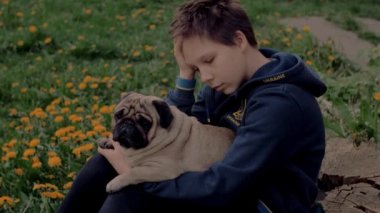 5 MAY, 2022 - WARSAW, POLAND: Sad Ukrainian refugee boy sitting in the yard full of dandelions and petting his dog. Teenager props his head up with his hand. Slow motion.