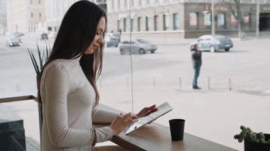 Young woman using a tablet and drinking coffee from the black paper cup sitting in cafe near the big window and watching city life. Handheld slow motion medium shot.