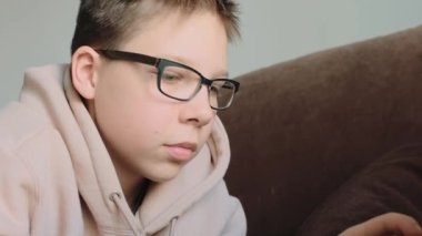 Caucasian teenage boy in hoodie with eyeglasses using laptop laying on the coach at home. Close-up shot.