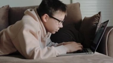 Caucasian teenage boy in hoodie with eyeglasses using laptop laying on the coach at home.