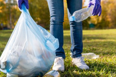 Bir kadın gönüllü, şehir parkında plastik çöp topluyor. Şişeyi çöp torbasına koyuyor. Ekoloji kavramı.
