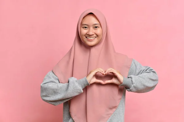 美しいアジアのイスラム教徒の女性は 愛を表現し 心の形をジェスチャーになり 私の愛 肯定的に笑顔 ヒジャーブを身に着けている ピンクの背景に対してポーズ ボディランゲージの概念 — ストック写真