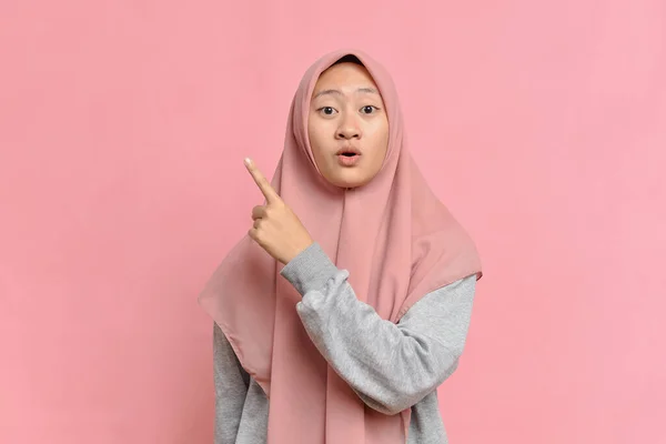 兴奋的亚洲美丽的年轻穆斯林女子的画像 指向一个复制空间 被粉红色背景隔离 — 图库照片