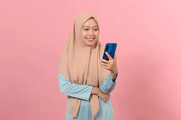 快乐微笑的年轻穆斯林女人手里拿着智能手机 看着屏幕发短信 用在线社交媒体应用程序孤立地站在粉色背景下 — 图库照片