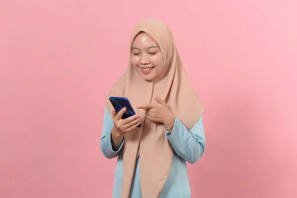 幸せな笑顔の若いイスラム教徒の女性はピンクの背景に孤立立ってオンラインソーシャルメディアアプリを使用して表示画面のテキストメッセージを見て 手にスマートフォンを保持します — ストック写真