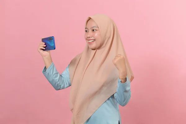 令人惊讶的是 年轻的亚洲穆斯林妇女戴着头巾 用智能手机庆祝一场以粉色为背景的胜利 — 图库照片