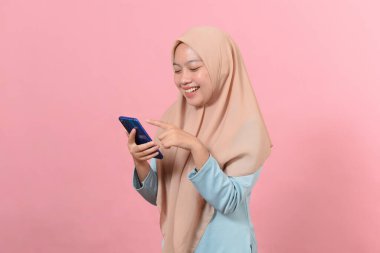 Mutlu gülümseyen genç Müslüman kadın elinde akıllı bir telefonla ekranda mesajlaşan mesajlara bakıyor pembe arka planda izole edilmiş duran sosyal medya uygulamalarını kullanıyor.