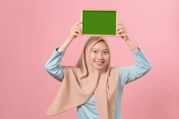 漂亮的年轻的穆斯林女人拿着粉红背景的黑板上的东西 — 图库照片