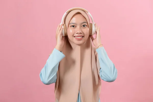 若い笑顔のイスラム教徒の女の子は 白いヘッドフォンで彼女の好きな音楽を聞きます カメラを見て 楽しさ 音楽愛好家 ピンクの背景に対して — ストック写真