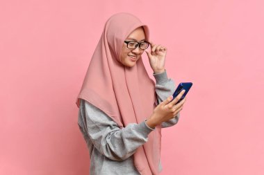 Güzel, çekici, genç Müslüman kadın. Akıllı telefona bak. Pembe arka planda izole edilmiş mesajı oku.