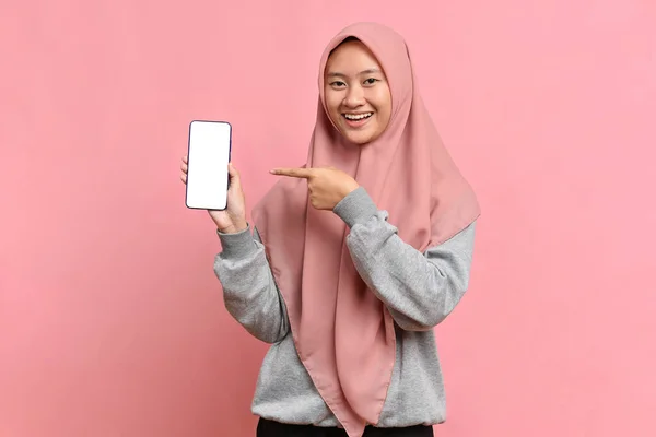 一个年轻迷人的穆斯林女孩 头戴头巾 面带微笑 手指指向一个粉色工作室背景的空白手机智能手机 文字的位置 有复制空间 — 图库照片