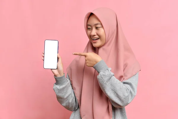 ヒジャーブを持つ若い魅力的なイスラム教徒の女の子の肖像画は簡単に笑顔とピンクのスタジオの背景に空白の携帯電話のスマートフォンで指を指します テキスト用の場所 コピースペース付き — ストック写真