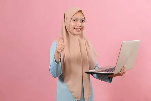 年轻美丽的快乐的穆斯林女子头戴头巾 手持笔记本电脑 用手指的大拇指来辨别粉色背景下的好迹象 — 图库照片