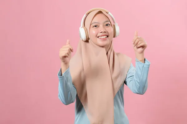 快乐的亚裔穆斯林女孩 一边用无线耳机听音乐 一边在粉色背景上跳舞 — 图库照片