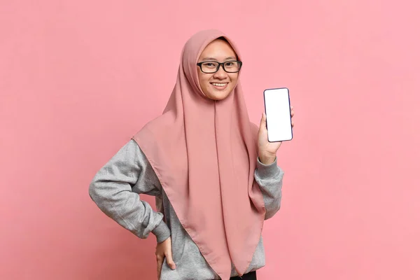 スカーフを着た若いアジアのイスラム教徒の女性はピンクの背景に隔離された画像やテキスト挿入のためのモックアップ画面と携帯電話を保持しています — ストック写真